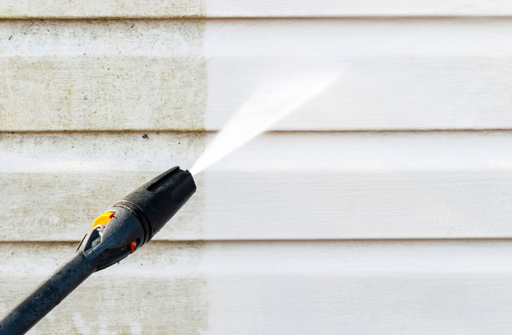 A pressure washer nozzle spraying water to clean the dirt off a white, horizontal siding of a house. The left side of the siding is dirty, while the right side is clean, showing a stark contrast between the two areas—an example of effective pressure washing Suffolk County services.
