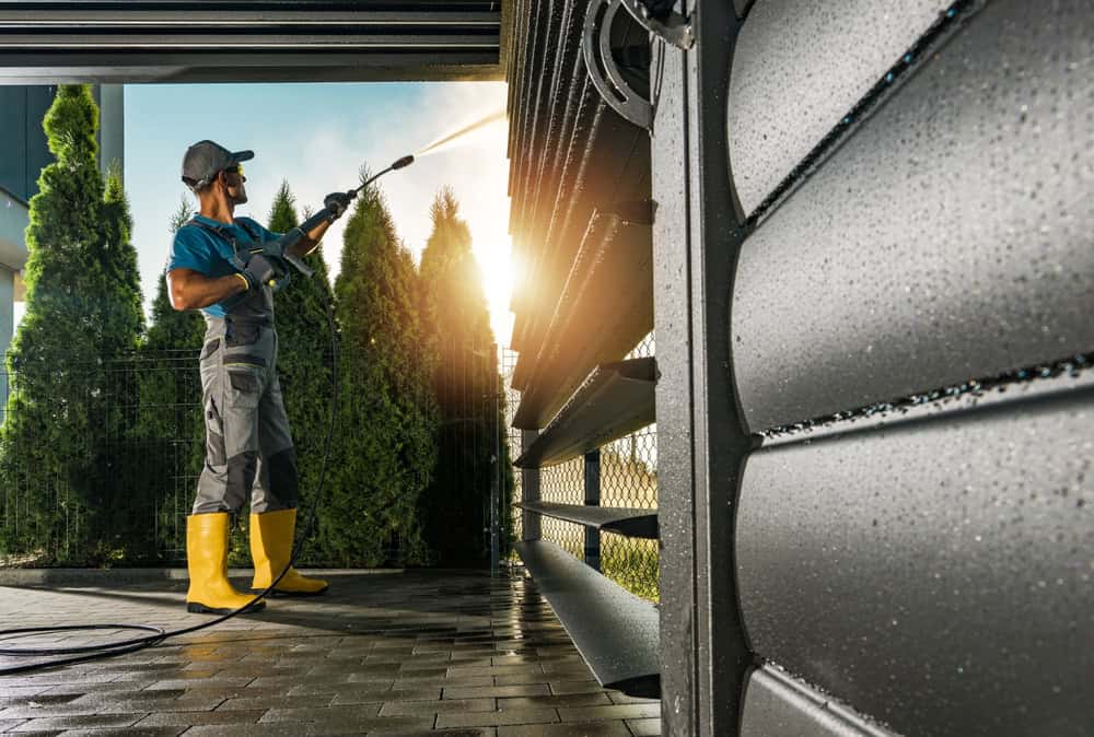 A person wearing gray overalls, a blue shirt, and yellow boots is power washing the exterior of a building. Tall green bushes are in the background, and the scene is lit by sunlight from behind. The wall and ground surface are wet—typical for effective Pressure Washing in Suffolk County.